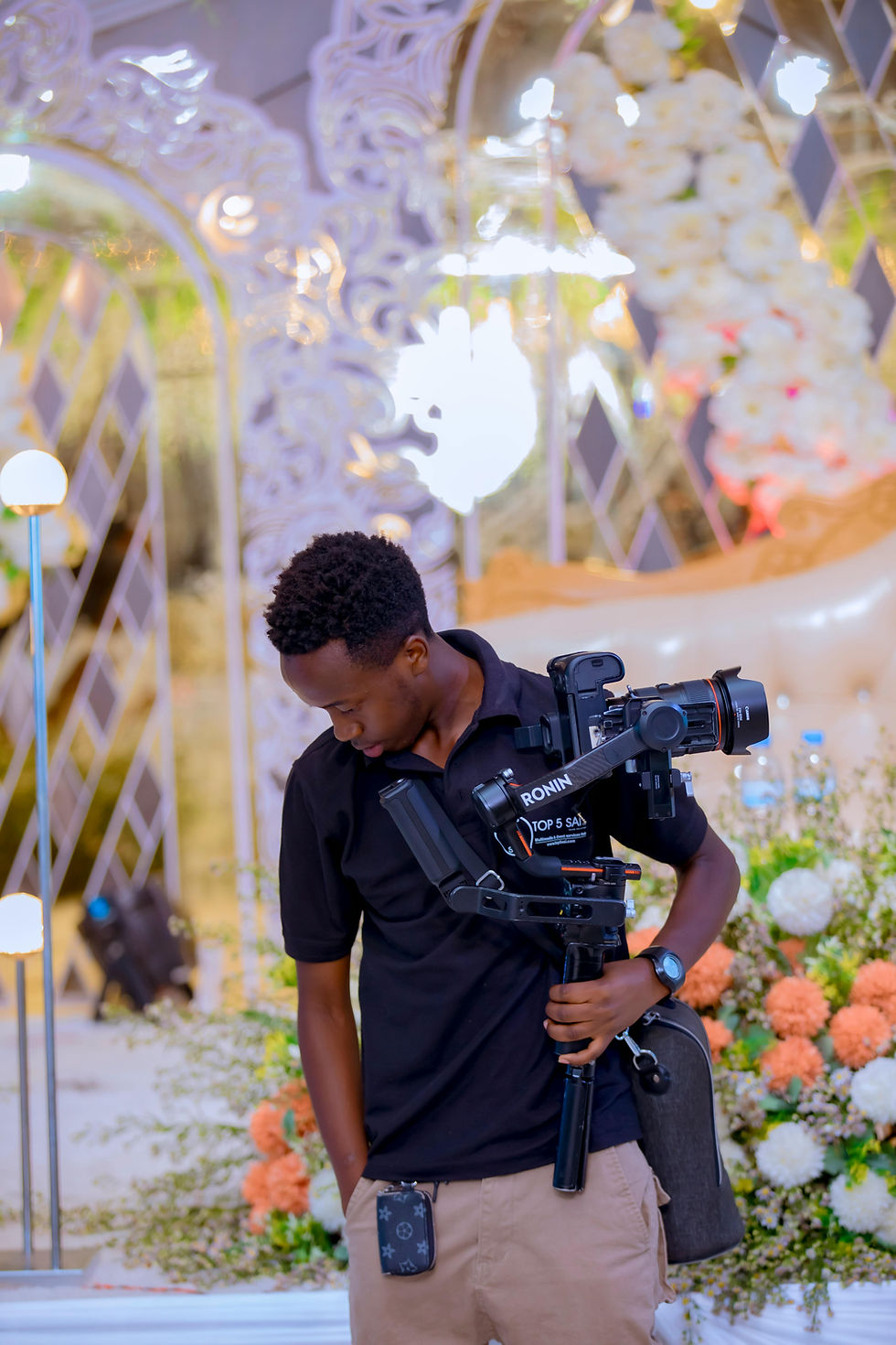 Young man holding a camera gimbal, looking down. Elegant floral decor in the background, with bright lights and ornate mirrors.