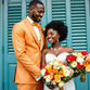 Bride in white dress and groom in orange suit smile at each other, holding vibrant rose bouquets. Blue shutters in the background.