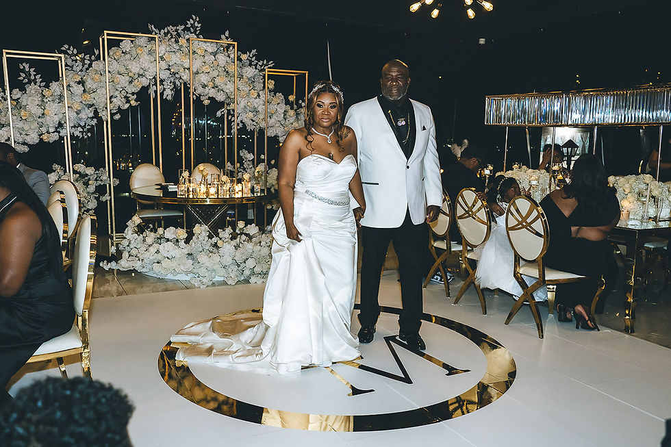 Bride and groom pose at a lavish white-and-gold wedding reception with floral arches and an M monogram on the floor.