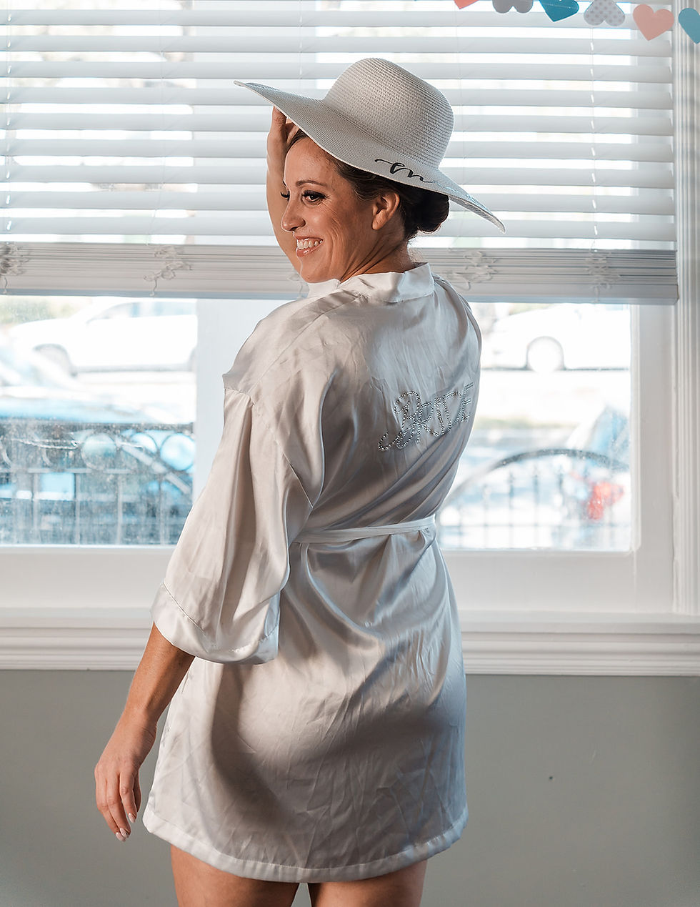 Woman in white robe and hat smiles near window blinds. Heart decorations hang above, and "bride" text is visible on her robe. Sunny day outside.