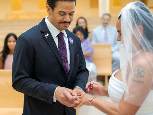 Groom places ring on bride's finger in a church. Both are smiling. Bride wears white dress and veil; groom, a dark suit with purple tie. New Orleans destination wedding photography