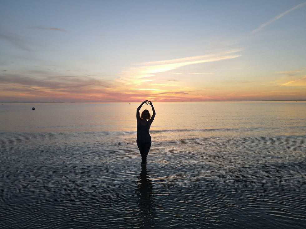 Frau steht im Meer und macht mit ihren Händen ein Herz, während die Sonne rotgelb aufgeht und das Meer in schöne Farben taucht.