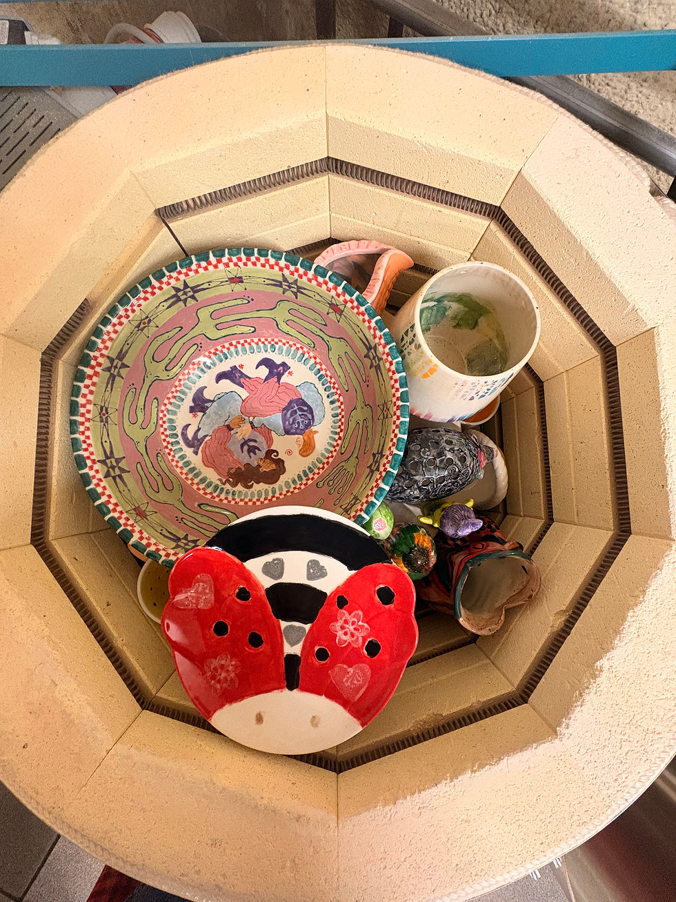 Colorful ceramic bowls and mugs stacked inside a beige kiln, including a ladybug-shaped piece and patterned bowl.