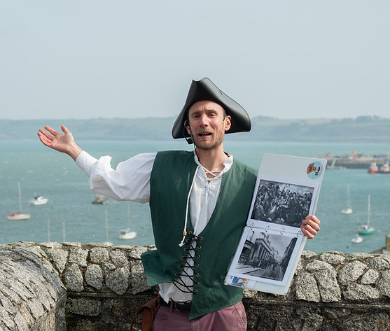 A pirate tour guide stands with his arms outstretched, a folder of historical images in his left hand. He is wearing a tricorn hat and a waistcoat. Behind him is Falmouth harbour; blue water dotted with little boats.