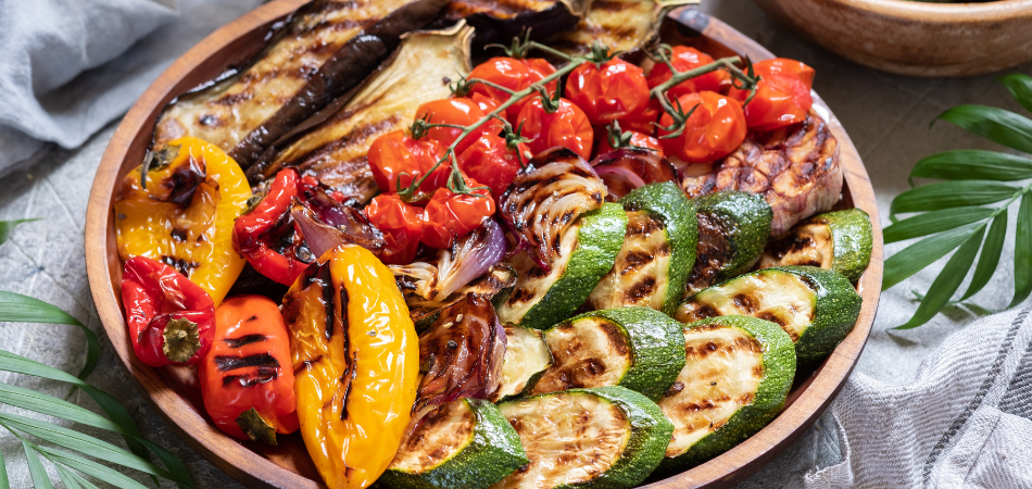 Colorful platter of grilled vegetables—zucchini, peppers, eggplant, tomatoes, and onions—on a rustic wooden dish.