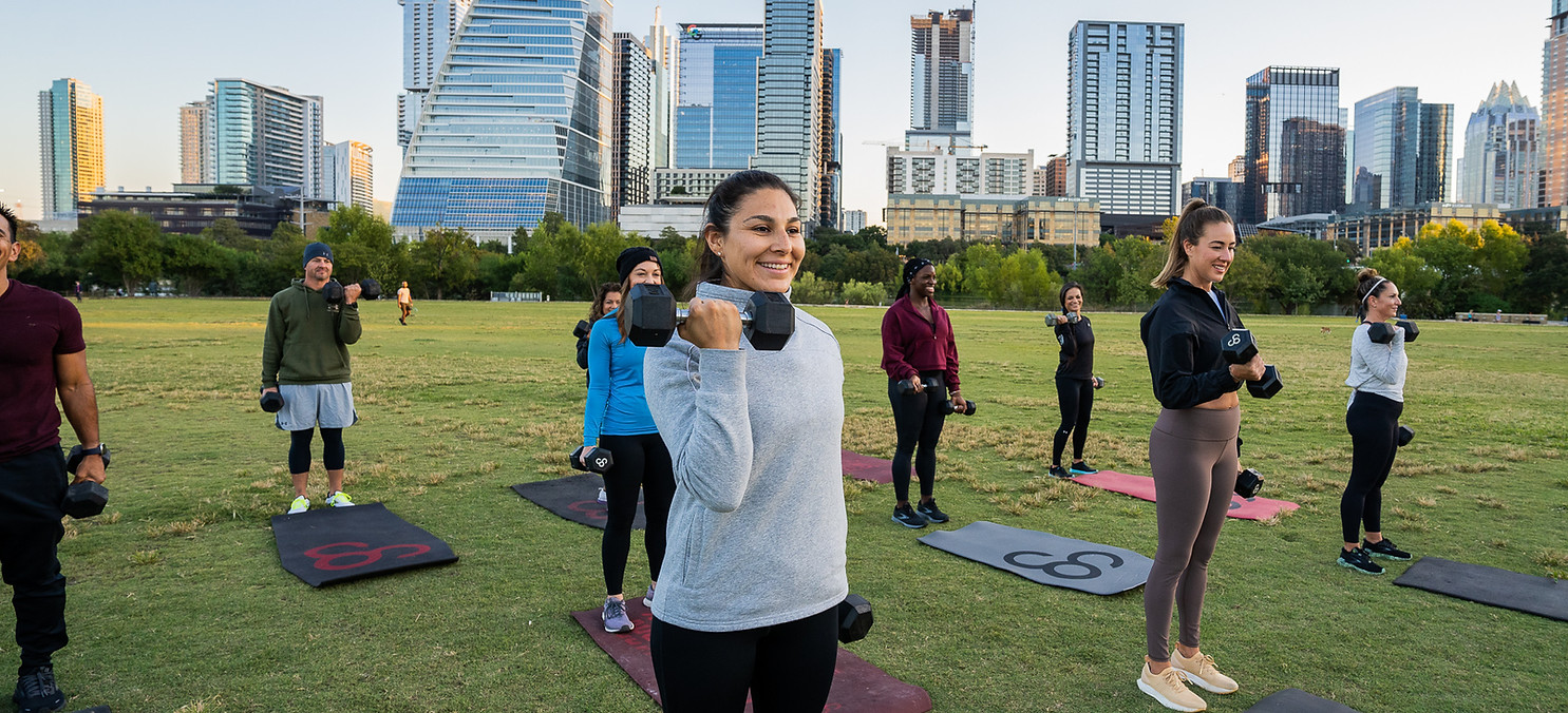 Group of people exercising with weights outdoors, Austin skyline in background. Camp Gladiator