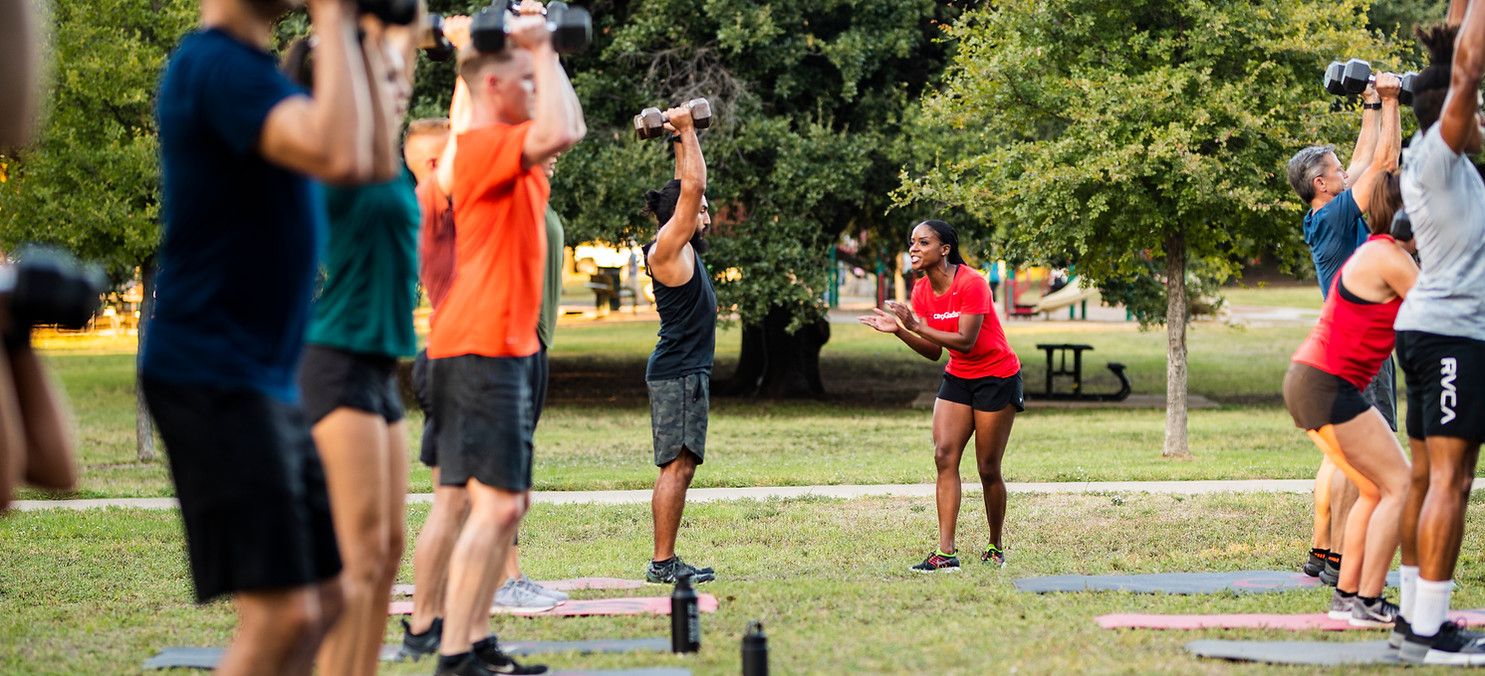 Group of people lifting weights outdoors with an instructor in the park.