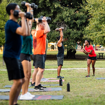Group fitness class lifting weights outdoors in a park Camp Gladiator.