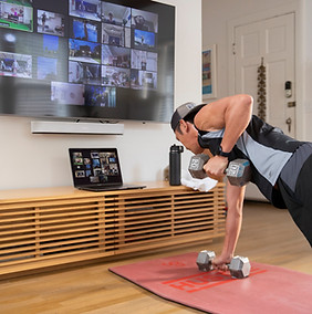 Man lifting dumbbells, watching online workout on laptop and TV screen. Camp Gladiator