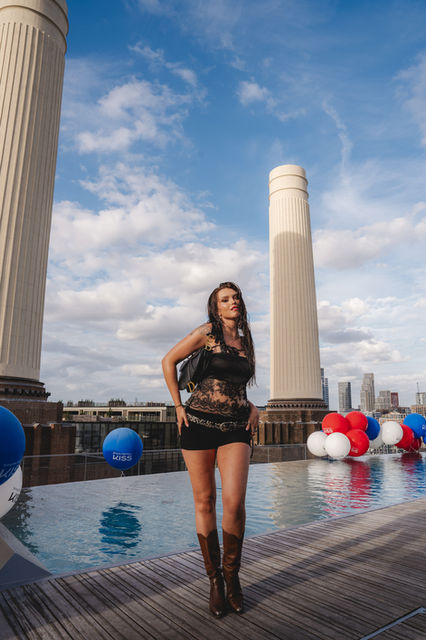 Photo taken by London event photographer Margot Jakobson at the Kiss Cosmetics Summer Party, JOIA Battersea. The image features a woman in a black lace top and skirt, posing confidently between Battersea's iconic chimneys with Kiss-branded balloons in the background.