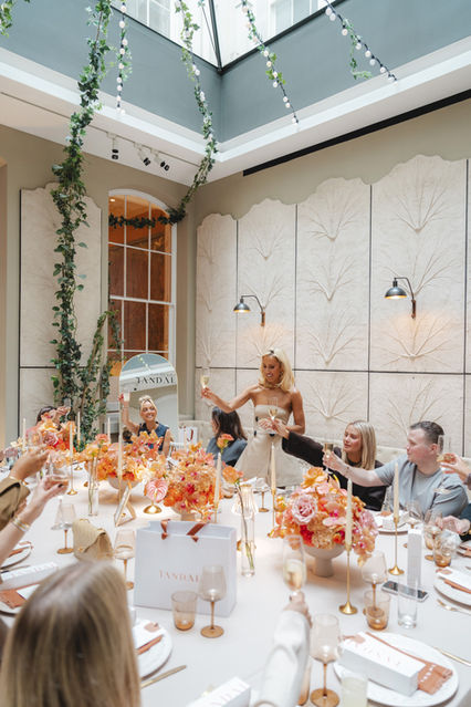 At the 'Tandae' event, Georgia Louise in a beige dress raises her glass in a toast at a lavishly set table adorned with peach-colored florals. Captured by London event photographer Margot Jakobson.