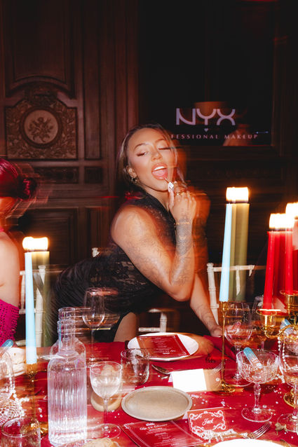 A woman in a black lace dress applies NYX Cosmetics lipstick at a lavish event table with candles and red decor. A bold and dynamic shot by London events photographer Margot Jakobson.
