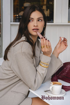 Gold Bohomoon jewellery styled with a beige blazer as the model applies lip balm in a café setting, photographed by Margot Jakobson in London.