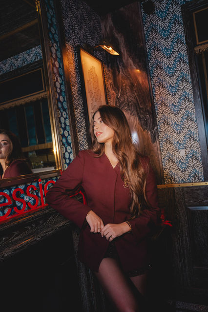 A stylish woman in a burgundy blazer poses confidently next to a mirror reflecting Essie branding in a luxurious venue. Photographed by London events photographer Margot Jakobson.