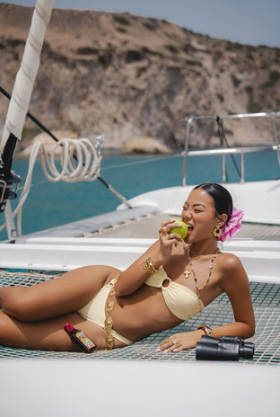 A stylish woman in a yellow bikini, wearing gold accessories, relaxes on a yacht and eats an apple, with scenic cliffs in the background, shot by fashion photographer Margot Jakobson for Selfie Leslie.