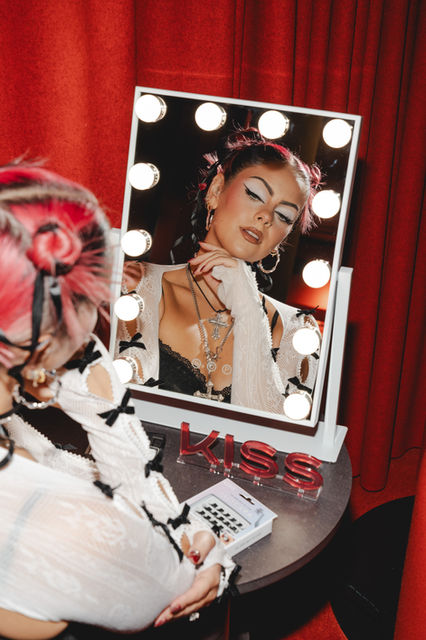 London event captured by Margot Jakobson features a woman with Kiss eyelashes, posing reflectively before a lit vanity mirror, exuding style and confidence.