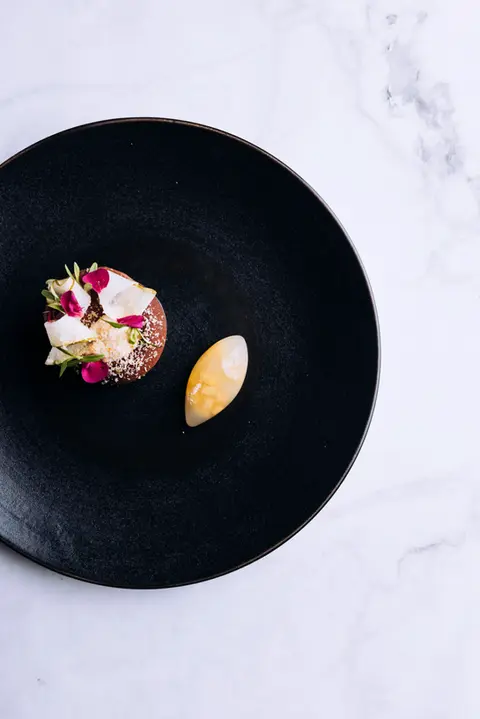 Elegant black plate featuring English pear mousse topped with 70% Ocoa dark chocolate, accompanied by smoked fudge, tarragon, and a toasted walnut anglaise, creating a sophisticated dessert experience.