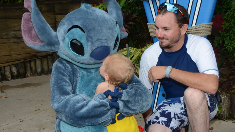 A little girl hugs Stitch as an adult male looks on with a smile