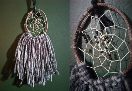 A small dreamcatcher made of a locally sourced branch bent into a oval. The dreamcatcher web has some black and metallic-silver beads. The bottom of the dreamcatcher circle is decorated with light and dark grey yarn.