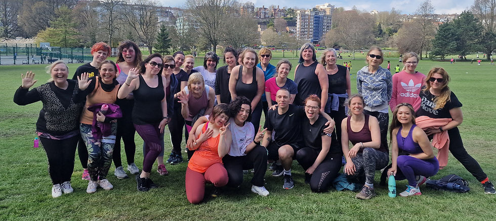 A big group of people smiling and posing in a Brighton park after a zumba class. Trees and buildings in the background. Some wave, showing a relaxed and cheerful mood. 
