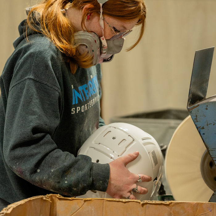 Woman in respirator and INTERSPORT hoodie polishing white helmet.