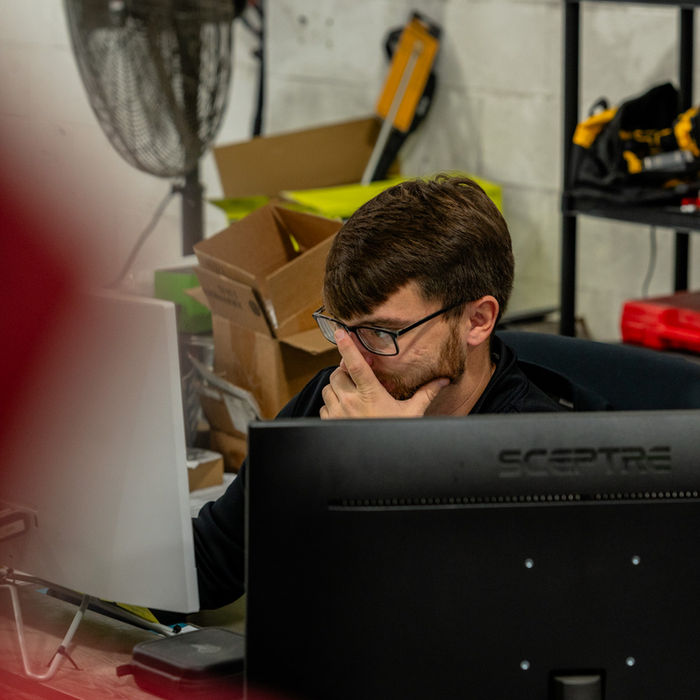 Focused man in glasses working on dual monitors in busy workshop.