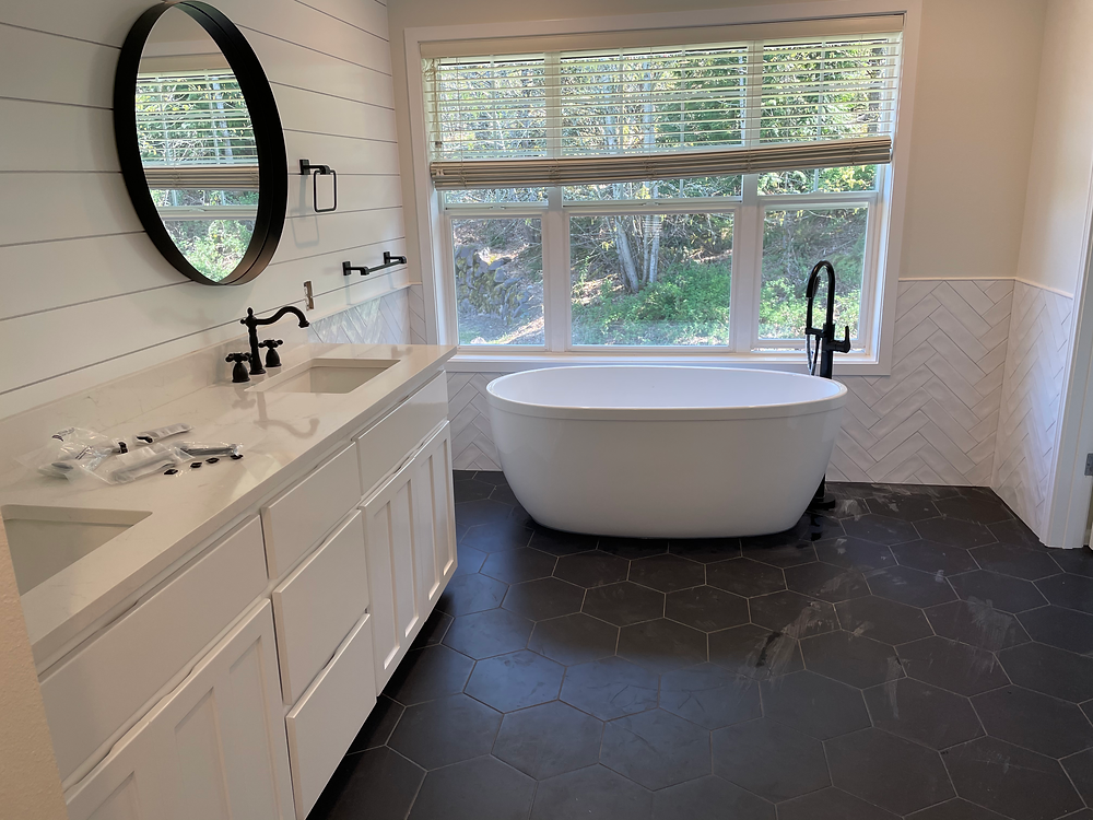 Modern black and white bathroom with freestanding tub, white vanity cabinets, large window, and dark tile flooring.