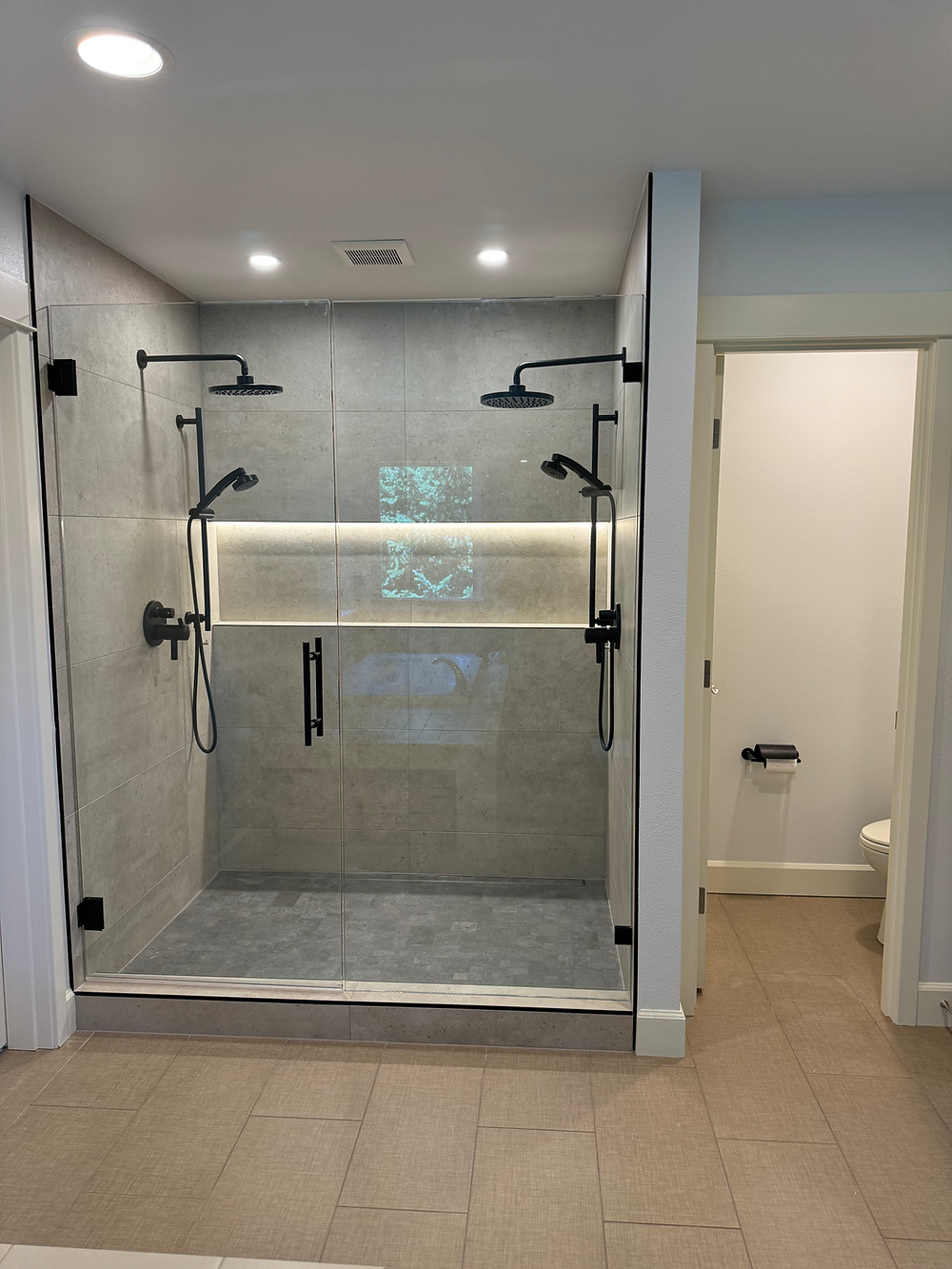 Modern bathroom shower remodel with frameless glass door, gray tile walls, and built-in illuminated shelving.