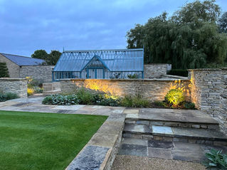 cotswold stone garden with large traditional green house lighting along low wall illuminating the bedding plants