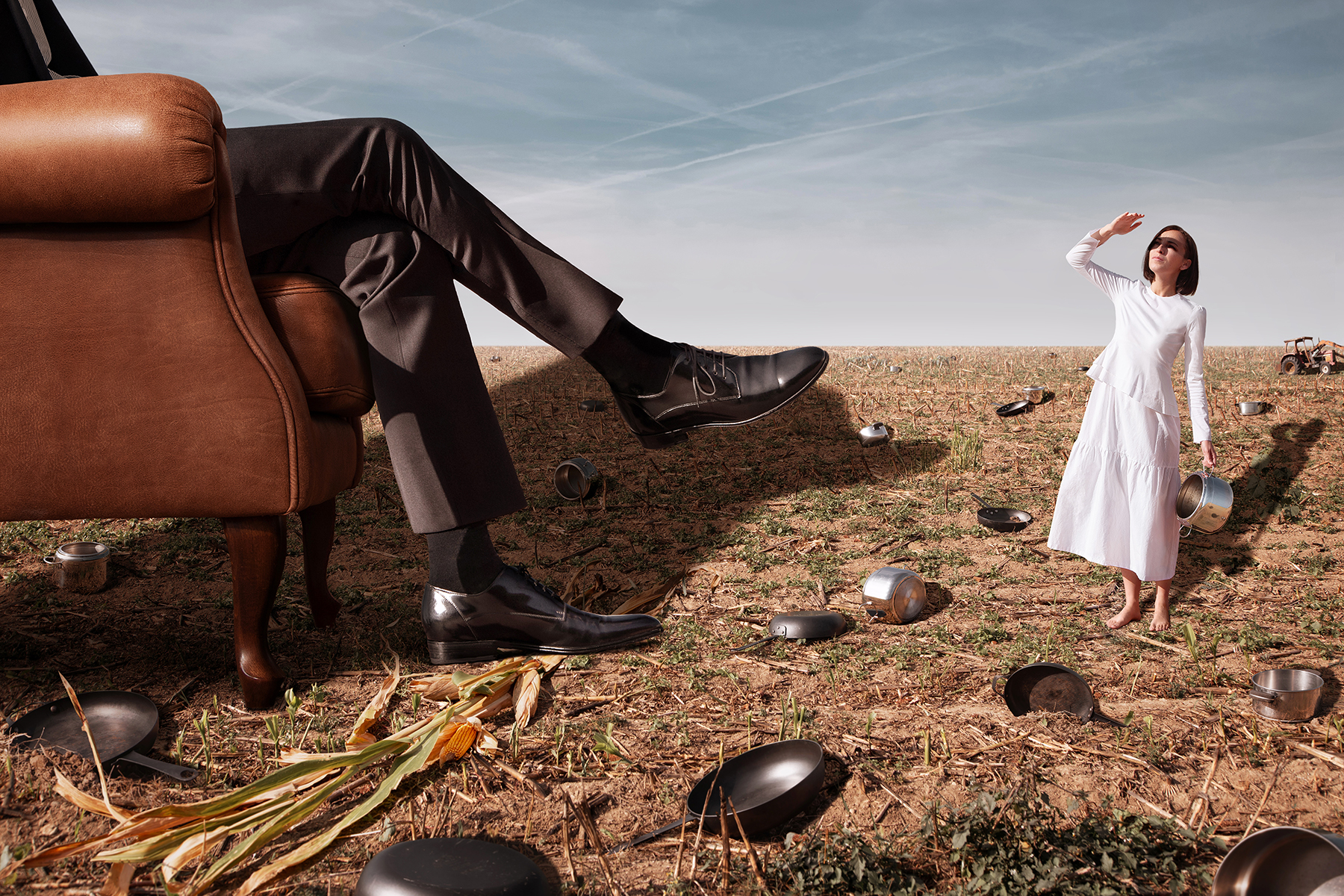 Surreal image about famine, domination and control. A girl with an empty pot on a dry field in front of huge crossed legs on an antique armchair.