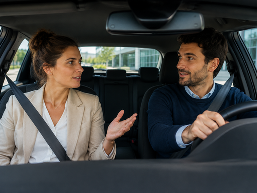 Deux collègues bouclés en voiture, discutant et souriant; la femme gesticule, devant un immeuble moderne.