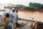 A man and two boys stand on an elevated roadside, looking out over floodwaters that have submerged the landscape.