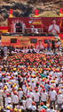 A sea of people in red and yellow caps is seen in a campaign stage. Fluttering flags and loudspeakers are seen. 