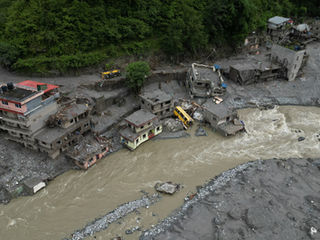 A gush of floodwater flows through the area, with homes buried under mud. A few houses remain on the other side, but most are surrounded by debris.