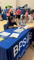 A Vietnamese volunteer wearing a navy T-shirt, leans across an information table to assist a man filling out paperwork at a community event. Families queue at nearby stalls while children wait beside their parents.