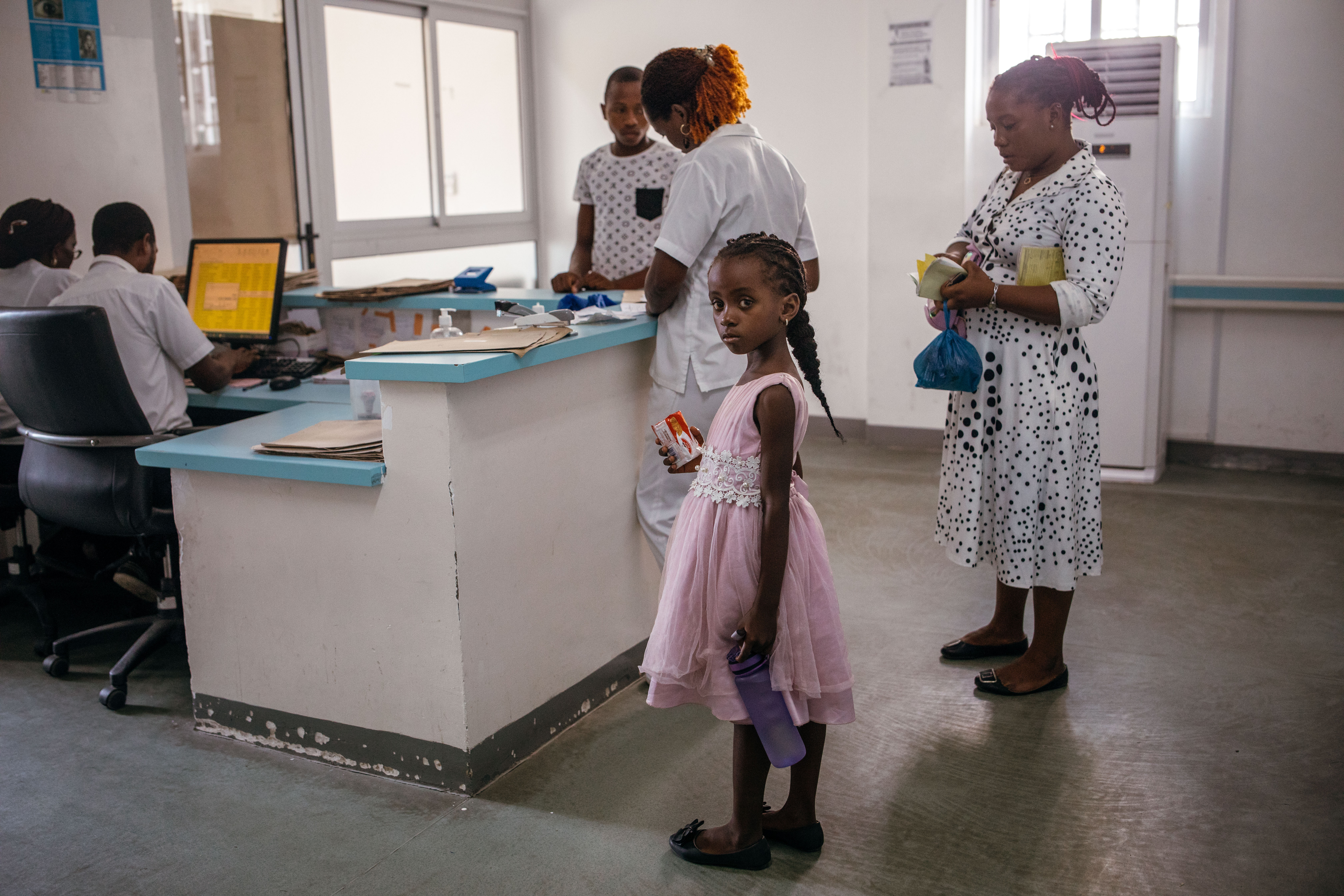 A young girl in a pink dress, her right hand holding medicine, stands at the hospital front desk. Behind her are three adults minding their own businesses, and at the table are two medical professionals working on a computer.