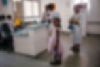 At the hospital reception, a young girl wearing a pink frock looks directly into the camera, while other people can be seen in the background.