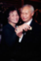 An older Asian man with a receding hairline holds his wife’s hand. She beams at the camera; both appear joyful.