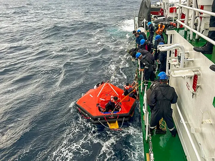 Rescue teams dressed in black assist survivors. An orange life raft is seen alongside as the crew members are pulled to safety.