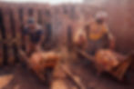 Two workers load bricks into wooden wheelbarrows inside a brick kiln. Stacks of baked bricks surround them as dust rises from the ground.