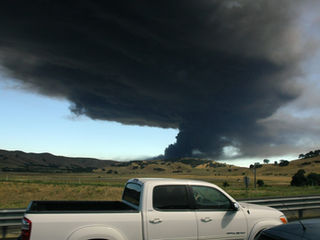 A vehicle drives down a highway with a dark cloud of black smoke engulfing the sky in the background.