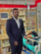 A Nepali doctor in a suit stands next to a patient lying on a hospital bed