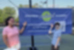 Two people wearing glasses smile and point at a "Southern Pickleball Academy" banner on a pickleball court, with a fence and trees behind.