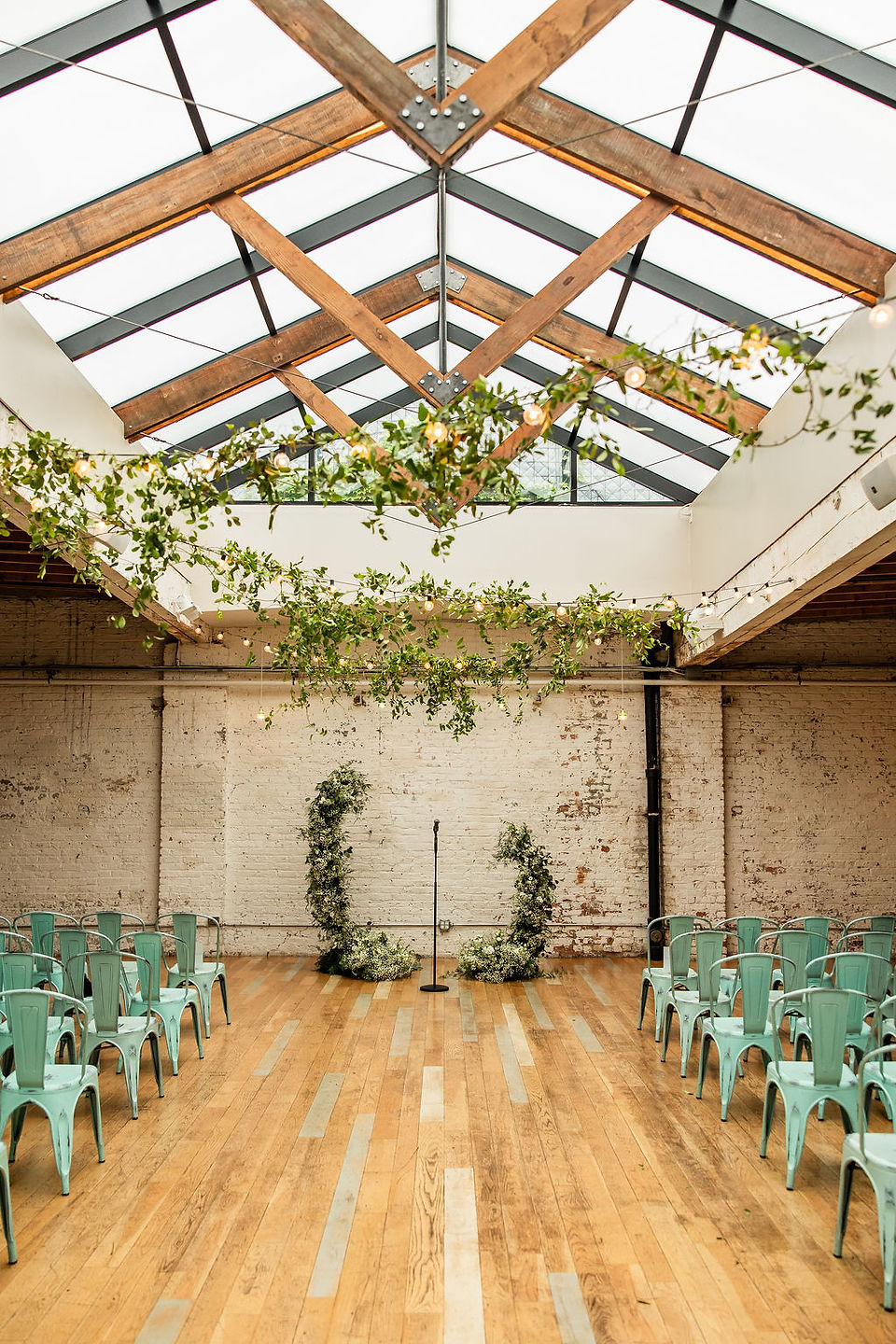 Wedding ceremony site with industrial brick wall, blue metal chairs, green foliage arch, skylight with wooden beams and foliage hanging at The Joinery 