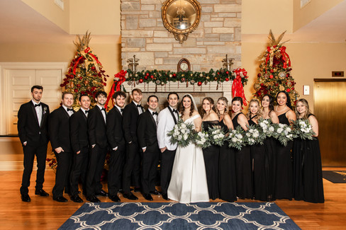 full wedding party with black bridesmaid dresses and black groomsmen tuxes standing in front of a Christmas decorated fireplace