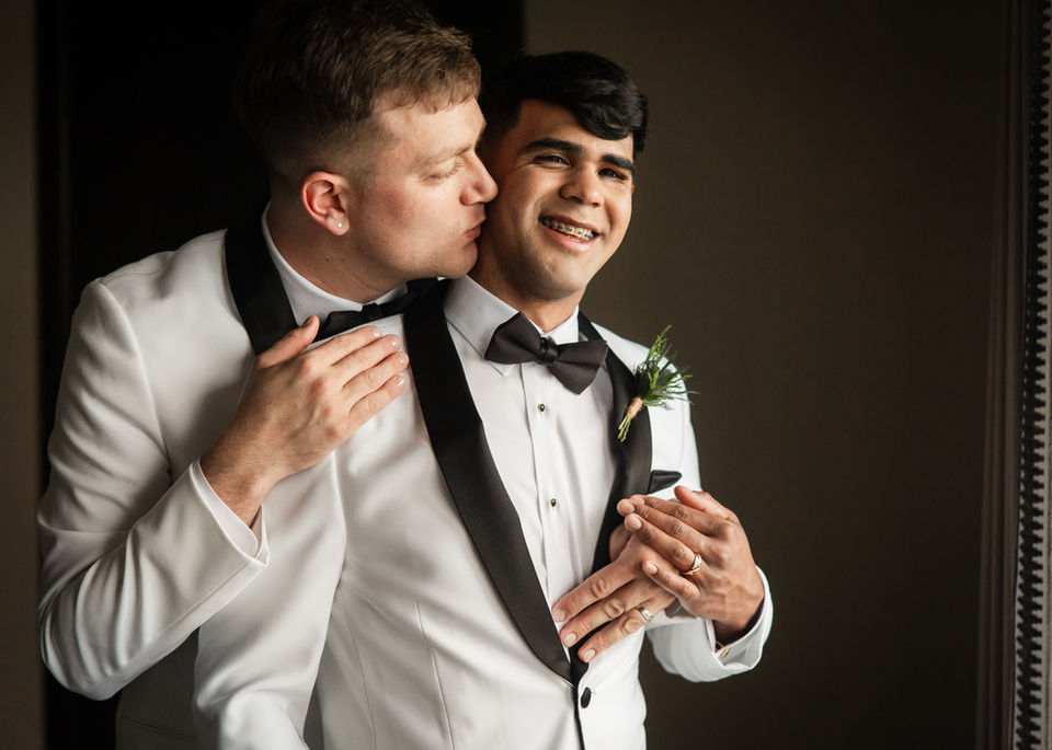 LGBTQ couple in white tuxes hugging by a window