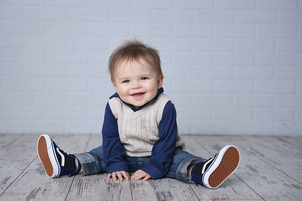 Baby boy in a cable-knit vest seated on a whitewashed wood floor against a white brick backdrop during a Sacramento sitter milestone session