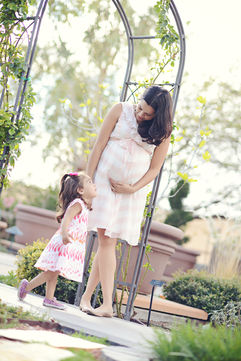 Pregnant mother in a pink-striped sundress smiling at her young daughter beside a garden trellis during a Sacramento maternity session