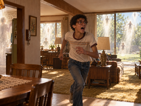 A frightened teenager wearing glasses races through a 1970s living room as glowing figures rise from the house and backyard.