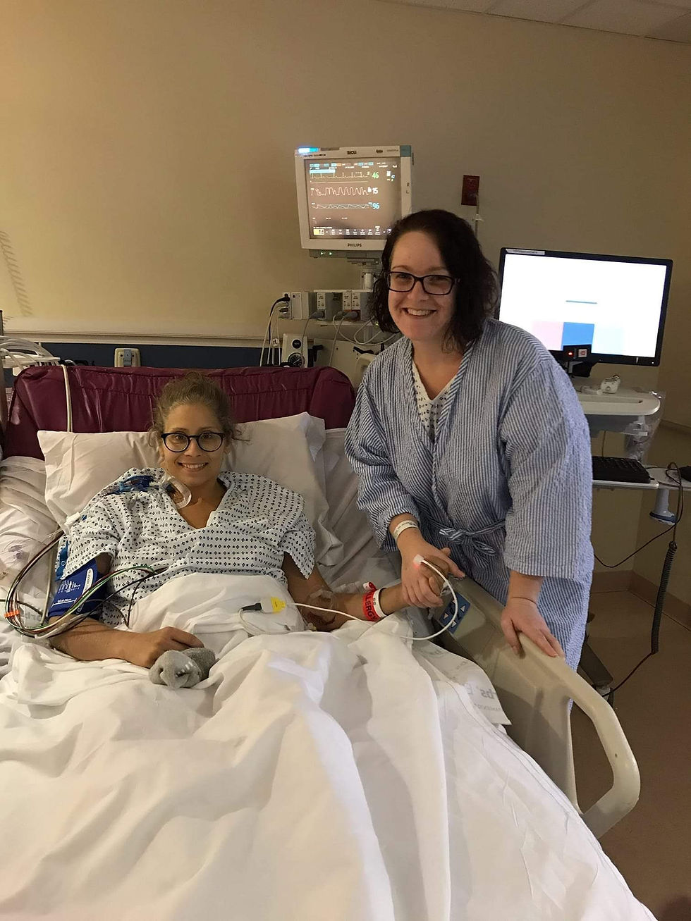 Two female friends in hspital gowns holding hands in a hospital room
